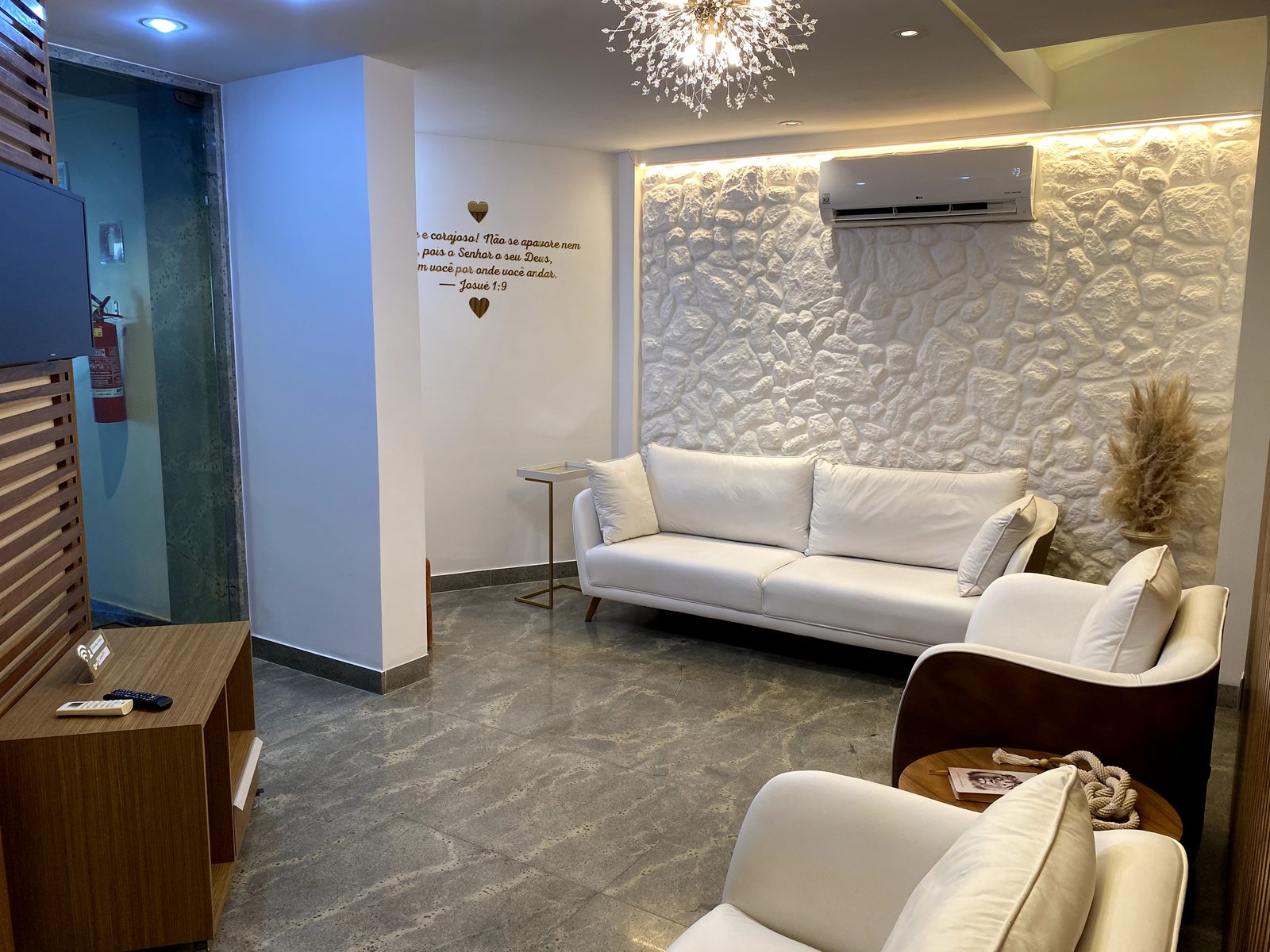 Sala de espera com sofás brancos, parede de pedra e lustre dourado